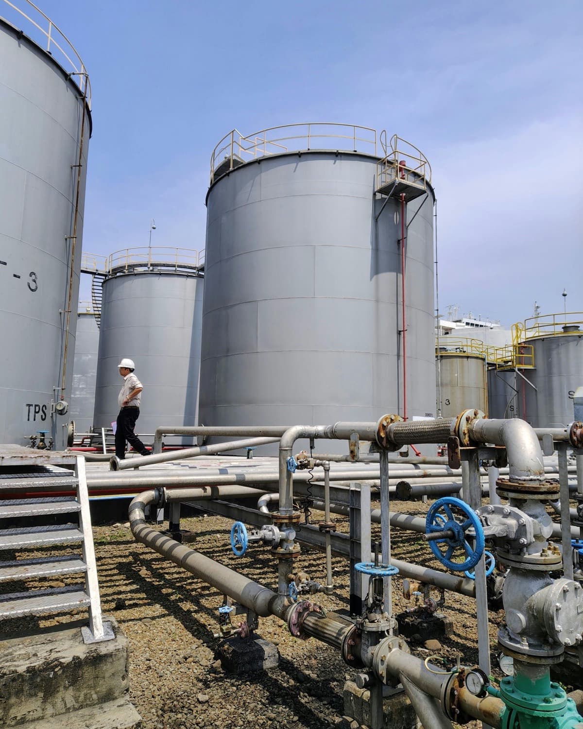 Engineer walking beside large industrial oil storage tanks and pipeline corridors under clear sky